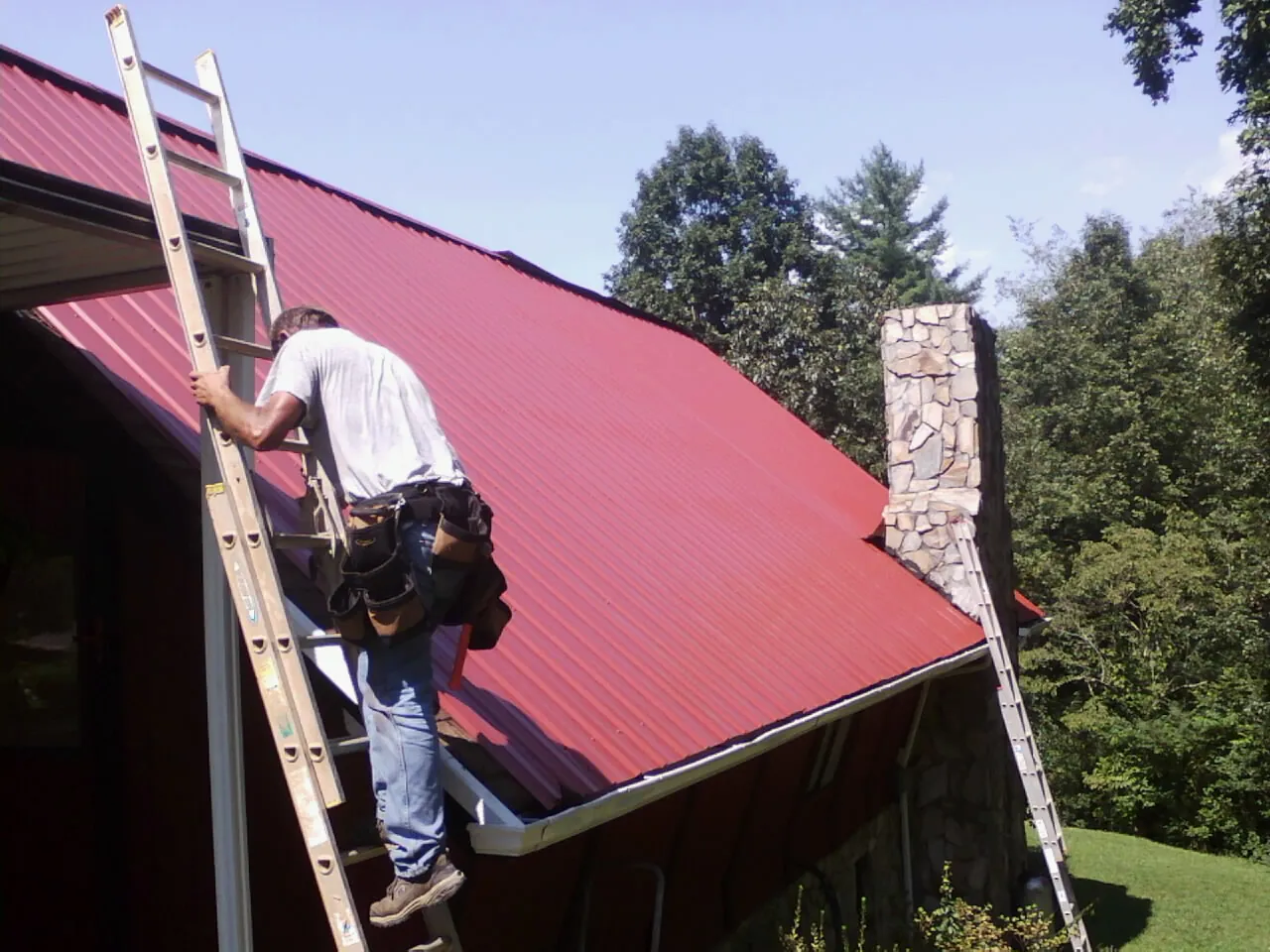 Roofer on a ladder beside a red metal roof and stone chimney — Soffit & Fascia Repair by Roofing Brothers in Elizabethport